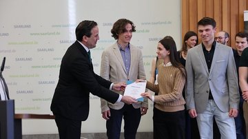 Drei Schüler stehen rechts und bekommen von einem Herrn im Anzug eine Urkunde überreicht