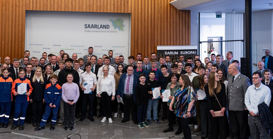 Ein großes Gruppenbild mit rund 70 Personen in verschiedenen Uniformen und Anzügen vor einer Wand mit dem Saarland-Logo