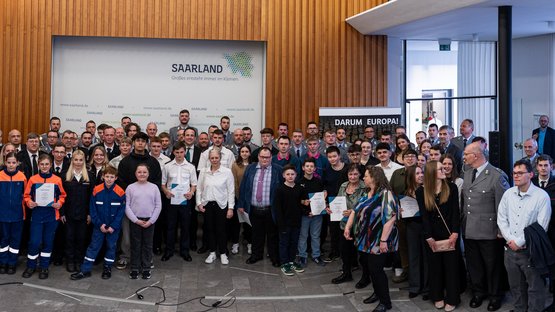 Ein großes Gruppenbild mit rund 70 Personen in verschiedenen Uniformen und Anzügen vor einer Wand mit dem Saarland-Logo