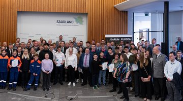 Ein großes Gruppenbild mit rund 70 Personen in verschiedenen Uniformen und Anzügen vor einer Wand mit dem Saarland-Logo