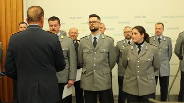 Drei uniformierte Soldaten erhalten eine Urkunde von einem Herrn, der der Kamera den Rücken zudreht