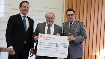 Ein uniformierter Herr und zwei Herren im Anzug halten einen Scheck in die Höhe
